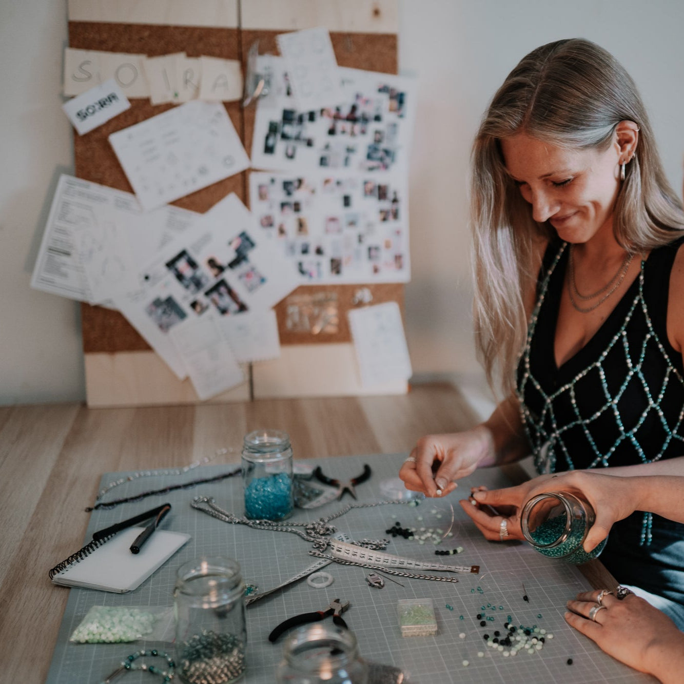 Sarah Hanisch (Gründerin von SOIRA) bei der Arbeit an Produkten. Sitzend am Tisch mit Perlen und Edelstahl Materialien. Im Hintergrund Inspirationsboards der Firma SOIRA